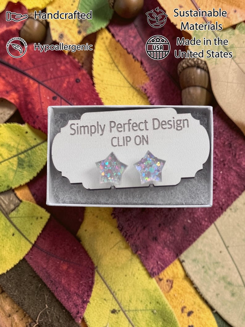 Op de afbeelding: Stervormige clip-on oorbellen met glitter, gepresenteerd in een kleine witte doos. De doos ligt op een bed van kleurrijke herfstbladeren. De tekst "Simply Perfect Design" en "CLIP ON" zijn zichtbaar op de doos.