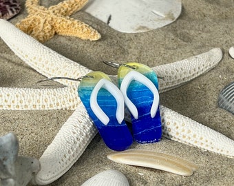 May include: A pair of dangle earrings shaped like flip-flops, featuring a gradient color pattern transitioning from yellow at the top to light blue and deep royal blue at the bottom, with white straps and silver-toned metal ear wires.