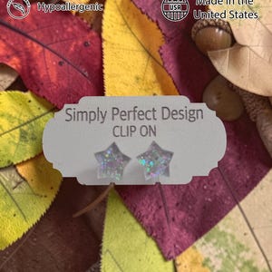 Op de afbeelding: Clip-on ster-oorbellen met iriserende glitter, gepresenteerd op een kaart met de tekst "Simply Perfect Design". De achtergrond toont herfstbladeren en eikels. De oorbellen zijn handgemaakt, hypoallergeen en gemaakt in de VS.