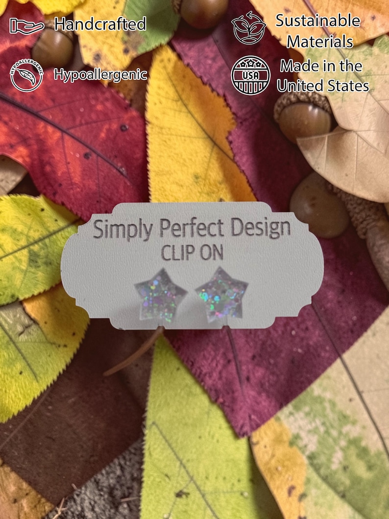 Op de afbeelding: Clip-on ster-oorbellen met iriserende glitter, gepresenteerd op een kaart met de tekst "Simply Perfect Design". De achtergrond toont herfstbladeren en eikels. De oorbellen zijn handgemaakt, hypoallergeen en gemaakt in de VS.