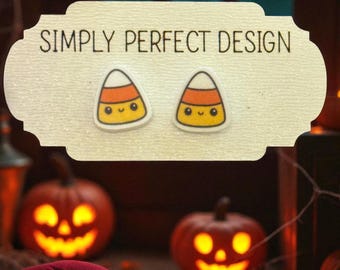May include: A pair of stud earrings shaped like candy corn with smiling faces, displayed on a cream-colored card with the text SIMPLY PERFECT DESIGN printed in black. The earrings feature white, orange, and yellow horizontal stripes.