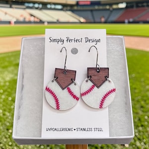 Könnte beinhalten: Ein Paar Ohrhänger mit einem weißen, glitzernden, kreisförmigen Baseball-Design mit roten Nähten, befestigt an einer dunkelbraunen, hölzernen Home-Plate-Form. Die Ohrringe hängen an Edelstahlhaken auf einer weißen Präsentationskarte mit dem Text Simply Perfect Design und Hypoallergenic Stainless Steel.