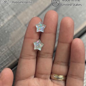 Op de afbeelding: Handgemaakte ster-vormige oorbellen met iriserende glitter. De oorbellen zijn hypoallergeen en gemaakt in de VS. De afbeelding bevat ook de tekst "Duurzame materialen".