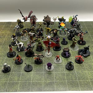 May include: A collection of fantasy miniature figurines, including dragons, demons, and humanoids, arranged on a green grid surface. The figurines are painted with various colors and details, showcasing a variety of characters and creatures.