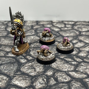 May include: Three small, painted, pink and white, skeletal figures with spindly legs and arms, standing on black bases. The figures are arranged in a triangle formation. A larger, painted, silver and gold figure with a sword is standing behind the three figures.