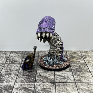 May include: A painted miniature figurine of a purple worm-like creature with sharp white teeth emerging from a rocky base, positioned next to a smaller painted miniature figurine of a bearded warrior holding a hammer and wearing a blue cloak.