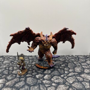 May include: A red and orange dragon miniature with purple accents and outstretched wings stands on a brown base. A smaller, silver miniature of a warrior stands in front of the dragon.