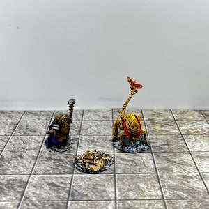 May include: A painted miniature figure of a bearded dwarf holding a hammer, a small pile of gold coins, and a yellow insectoid creature with a long tail and red accents, all standing on individual circular bases.