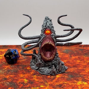 May include: A gray, tentacled monster figurine with a large open mouth, a single orange eye, and multiple tentacles. A blue and black twenty-sided die is to the left of the monster. The figurine is on a red and orange surface.