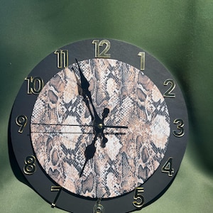 May include: A round wall clock with a snakeskin pattern in the center. The clock has a black frame and gold-colored numerals. The hands are black. The clock is a decorative item.