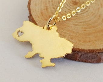Könnte beinhalten: Eine goldene Halskette mit einem Anhänger in Form der Umrisse eines Landes. Der Anhänger hat ein kleines Herz ausgeschnitten. Die Halskette liegt auf einer Holzoberfläche.