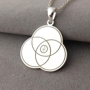 May include: A silver pendant necklace featuring a circular design composed of three overlapping circles. The pendant has a small, detailed design in the center and hangs from a delicate silver chain. The necklace is displayed on a gray surface.
