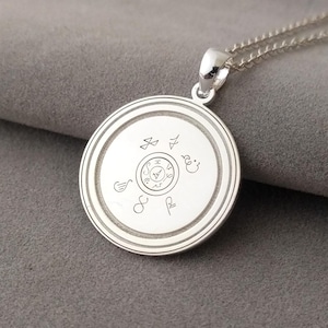 May include: A silver-colored pendant necklace with a circular pendant. The pendant features an engraved design with symbols and a central circular emblem. The necklace chain is also silver. The pendant is displayed on a gray surface.