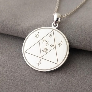 May include: A silver pendant necklace featuring a circular pendant with etched geometric symbols and a triangle design. The pendant hangs from a delicate silver chain. The necklace is displayed on a gray surface.