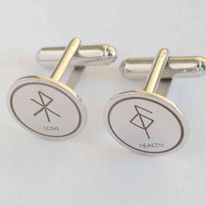 May include: A pair of silver cufflinks with circular engraved designs. One cufflink features a symbol and the word "LOVE". The other cufflink features a different symbol and the word "HEALTH".