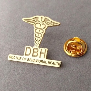 May include: Gold-colored pin with a medical caduceus symbol and the text "DBH Doctor of Behavioral Health".