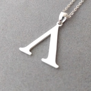 Puede incluir: Collar con colgante de plata con la letra griega Lambda. El colgante de plata pulida cuelga de una fina cadena. El fondo es gris neutro.