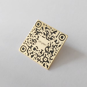 May include: A gold-coloured metal square with a black and white QR code and the text "stryker" printed on it.