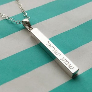 Peut inclure: Un collier en argent avec un pendentif rectangulaire long gravé de texte hébreu. Le pendentif est suspendu à une délicate chaîne en argent.