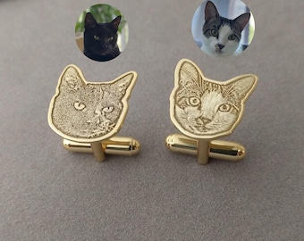 Puede incluir: Gemelos dorados con diseños de caras de gato. Los gemelos tienen una barra rectangular y un diseño de cabeza de gato. Dos imágenes circulares de gatos están encima de los gemelos. Los gemelos están sobre una superficie gris.