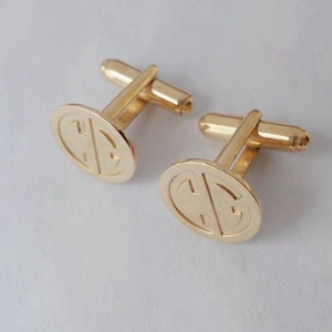 May include: A pair of gold cufflinks with a circular design featuring a monogram. The cufflinks have a simple, classic design.