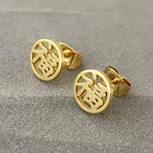Può includere: Un paio di orecchini a bottone color oro. Ogni orecchino presenta un design circolare con un carattere cinese ritagliato. Gli orecchini sono esposti su una superficie grigia.