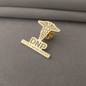 Può includere: Una spilla in metallo color oro con il simbolo del Caduceo e le lettere "DNP" con le parole "DOCTOR OF NURSING PRACTICE" sottostanti. La spilla è progettata per un dottore in pratica infermieristica.