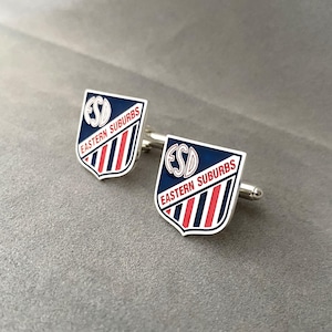 May include: A pair of silver cufflinks with a shield design. Each shield is navy blue with the letters "ESD" and the words "EASTERN SUBURBS" in white. Red and white stripes are on the lower half of the shield.