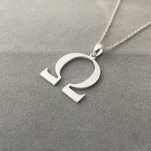 May include: A silver-colored pendant necklace featuring the Greek letter Omega. The pendant is a stylized, open design with a curved top and two straight lines at the bottom. The necklace chain is a delicate, silver-colored cable chain.