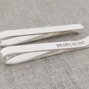 May include: Two silver tie clips. One clip has the phrase "you carry my heart" engraved on it. The clips are on a grey, textured surface. The clips are made of a shiny metal.