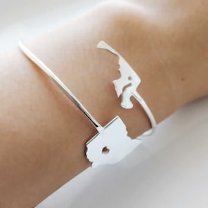 Peut inclure: Un bracelet manchette argenté avec un motif découpé représentant une carte d'État. Le bracelet est porté sur un poignet et présente une esthétique minimaliste et moderne. Le design ouvert facilite le port et ajoute une touche d'élégance.