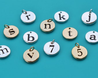 Puede incluir: Una colección de dijes redondos de metal plateado y dorado con letras y números. Los dijes tienen un agujero en la parte superior para sujetarlos a un collar o pulsera.