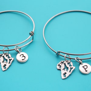 Puede incluir: Dos pulseras de brazalete expandibles de plata con dijes. Un dije es un recorte de plata del continente africano con una jirafa y otros animales. El otro dije es un círculo de plata con la letra "P" en él. La otra pulsera tiene un recorte de plata del continente africano con una jirafa y otros animales y un círculo de plata con el número "3" en él.