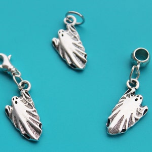 Op de afbeelding: Drie zilverkleurige spookbedels op een turquoise achtergrond. Elke bedel heeft een gestileerd spookontwerp met uitgesneden ogen en mond. Eén bedel heeft een karabijnhaak, een andere heeft een lus.