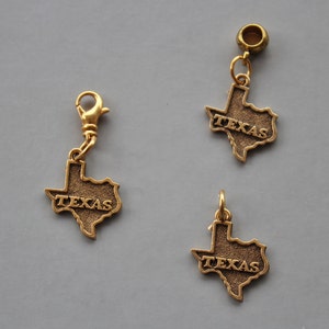 May include: Three gold-colored charms in the shape of the state of Texas. Each charm has the word "TEXAS" inscribed on it. One charm has a lobster clasp, one has a bead, and one has a loop.
