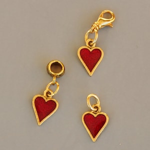 Pode incluir: Três pingentes de coração de ouro com esmalte vermelho. Os pingentes são de tamanhos diferentes e têm diferentes estilos de fixação.