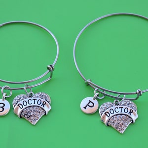 Peut inclure: Deux bracelets extensibles en argent avec des breloques en forme de cœur. Un charme a le mot "DOCTOR" avec des strass et la lettre "P" sur un charme rond. L'autre charme a le mot "DOCTOR" avec des strass et le chiffre "3" sur un charme rond.