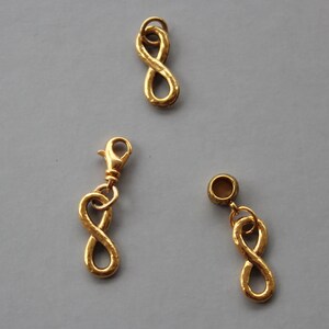 Pode incluir: Três pingentes de ouro com símbolo de infinito. Dois dos pingentes têm um laço na parte superior para prender a um bracelete ou colar. O terceiro pingente tem um fecho.