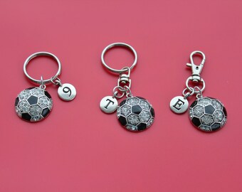 May include: Three silver keychains with soccer ball charms. The charms are black and white with rhinestones. Each keychain has a small silver charm with a letter or number. The charms read "E", "T", and "9".