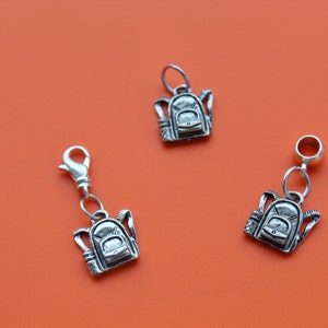 May include: Three silver charm pendants in the shape of a backpack. The charms are on a bright orange background.