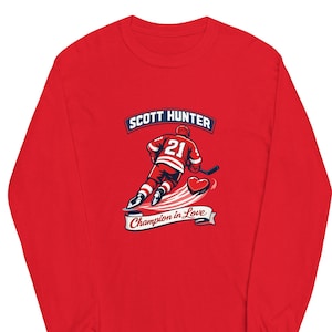 May include: A long-sleeve red shirt featuring a hockey player graphic. The graphic includes the name "SCOTT HUNTER" above a hockey player wearing the number 21, a heart, and the text "Champion in Love".