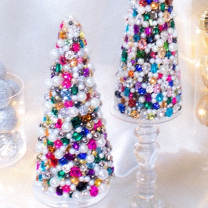 May include: Two small, cone-shaped Christmas trees decorated with colorful jewels, pearls, and rhinestones. The trees are on clear glass stands.