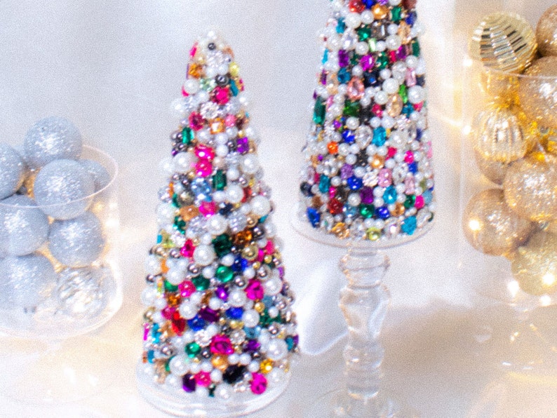 May include: Two small, cone-shaped Christmas trees decorated with colorful jewels, pearls, and rhinestones. The trees are on clear glass stands.