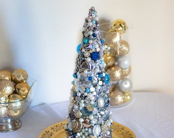 May include: A small, cone-shaped Christmas tree decorated with blue, white, and silver beads, buttons, and other embellishments. The tree is sitting on a gold plate.