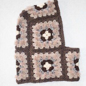 Peut inclure: Une cagoule au crochet avec un motif de carrés granny. La cagoule présente des carrés dans les tons marron, beige et gris. La cagoule est conçue pour couvrir la tête et le cou, avec une ouverture pour le visage.