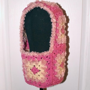 Peut inclure: Une cagoule rose et crème au crochet. La cagoule présente un motif carré avec une bordure duveteuse autour de l'ouverture du visage et du haut. The Knit Covenant est imprimé en haut à gauche de l'image.