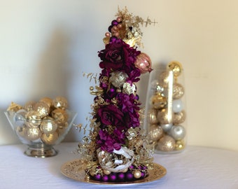 May include: A purple and gold Christmas tree centerpiece with a variety of ornaments and flowers. The tree is on a gold plate and surrounded by gold and white ornaments.
