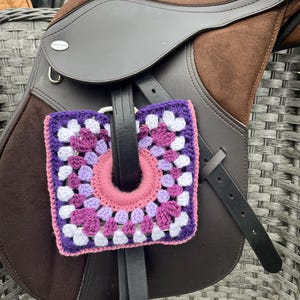 May include: A brown leather horse saddle with a square, crocheted pad. The pad features a granny square design in shades of purple, pink, and white, with a pink center. The saddle is on a gray wicker chair.