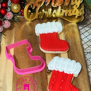 May include: A wooden board features a pink stocking-shaped cookie cutter and two decorated Christmas stocking biscuits. The biscuits are red with white icing and have a cable knit design. A gold "Merry Christmas" sign is in the background.