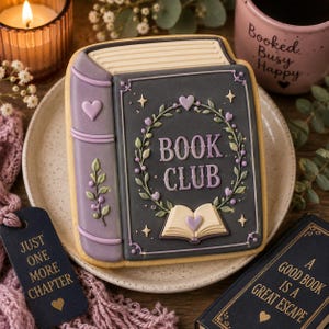 Puede incluir: Una galleta de azúcar rectangular decorada como un libro con cubierta gris oscuro, lomo morado y el texto "BOOK CLUB" en el centro. El diseño presenta una corona floral, pequeñas estrellas y un icono de libro abierto con un corazón morado.